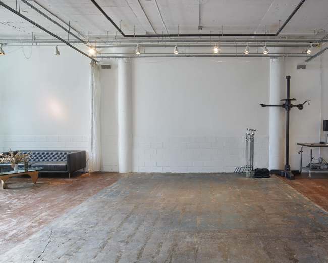 The image shows an empty industrial space featuring a black couch, a bare wall, and minimal furnishings, including a desk with a monitor and a clothing rack.