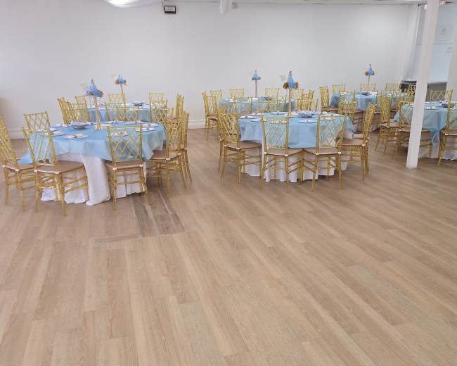 The image shows a spacious banquet hall set up with multiple round tables, each covered with blue tablecloths and surrounded by gold chairs.