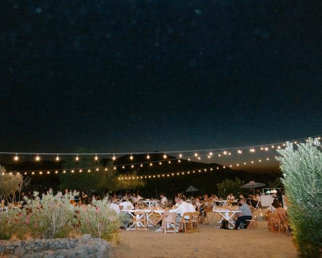 A garden event is set up outdoors at night, featuring tables surrounded by string lights and greenery.