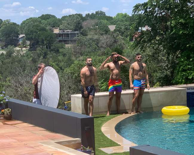 Four men in swimwear stand by a pool, while another man holds a reflector nearby.