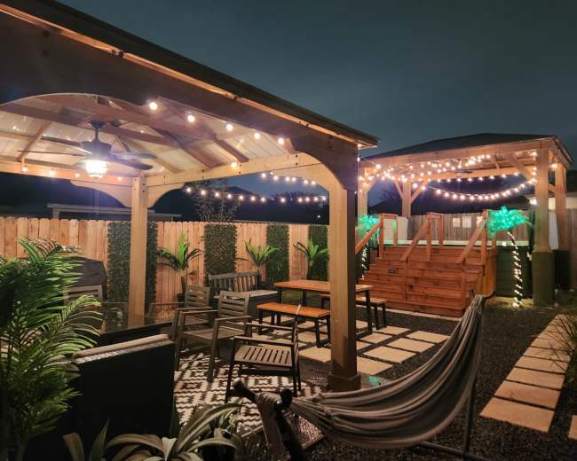 The image features a spacious backyard with a wooden gazebo, string lights overhead, seating areas, and a staircase leading to a raised deck.