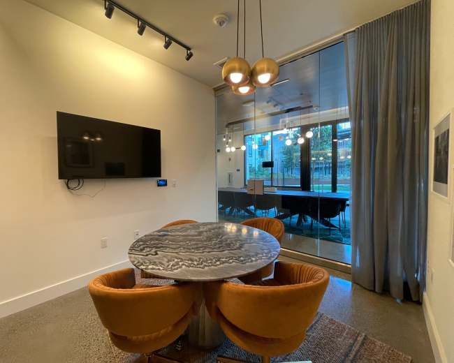 A modern meeting room features a round table with orange chairs, a large television mounted on the wall, and glass doors leading to an outdoor area.