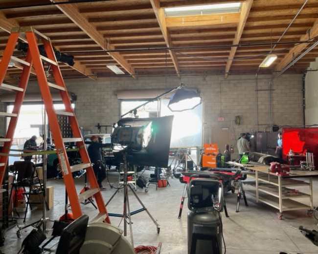 A workshop space with various filming equipment, tools, and a ladder, illuminated by natural light coming through a window.