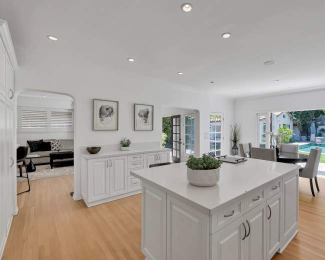 A bright kitchen with white cabinets and an island opens into a spacious living area featuring large windows overlooking a pool and outdoor seating area.