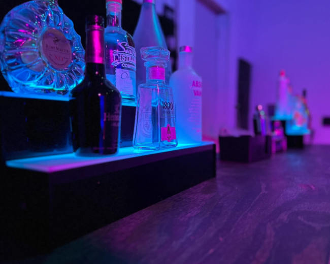 A variety of liquor bottles are arranged on a bar with colorful lighting.