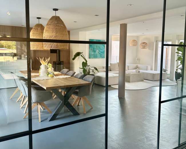 A modern open-concept dining area features a large wooden table, comfortable seating, and pendant lighting, with a view from an adjacent room through a glass partition.