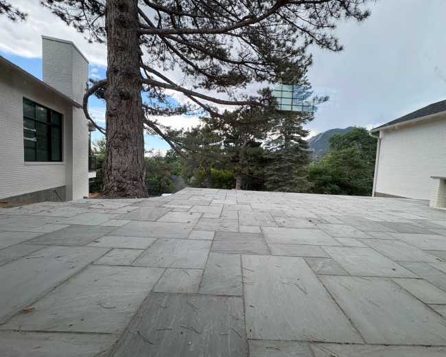 The image shows a spacious stone patio surrounded by trees and two white buildings.
