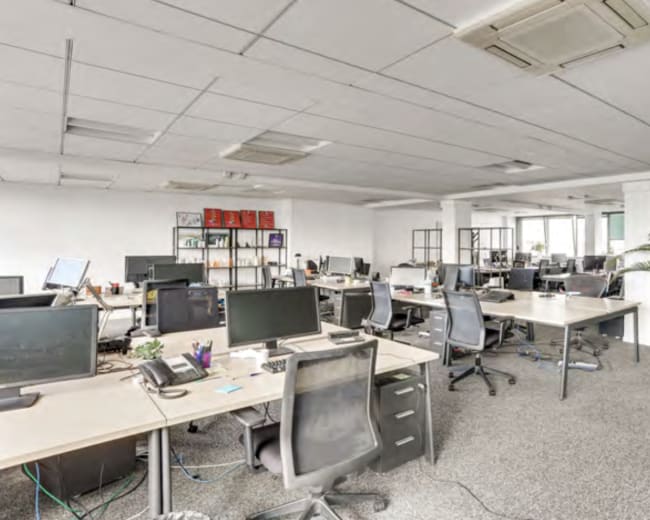 An open office space features multiple desks with computers, chairs, and decorative plants, with bright lighting and a minimalist design.