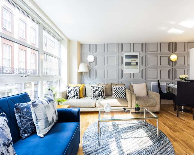 A modern living room features a sofa, blue chairs, a glass coffee table, and a dining area with a backdrop of patterned walls and large windows allowing natural light.
