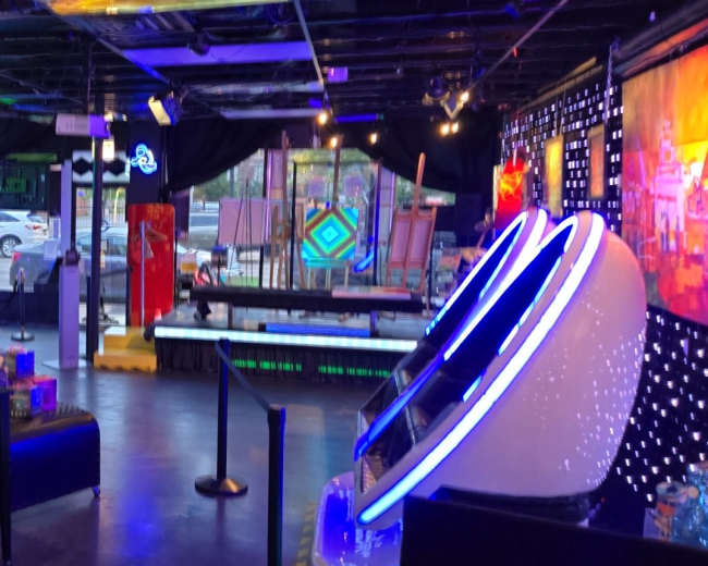 The image shows a colorful, modern arcade space filled with various gaming machines and art pieces, with vibrant lighting and decorative elements.