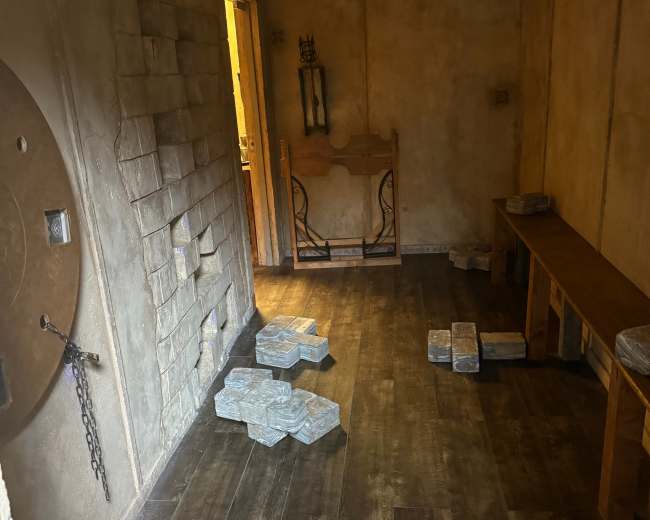 The image shows an empty room with textured walls, a wooden bench, scattered stone-like pieces on the floor, and a doorway leading to another area.
