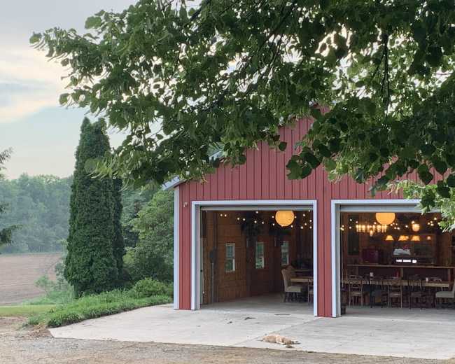 A red barn with open doors reveals a furnished interior, while a dog lies on the ground nearby amidst a view of fields and trees.