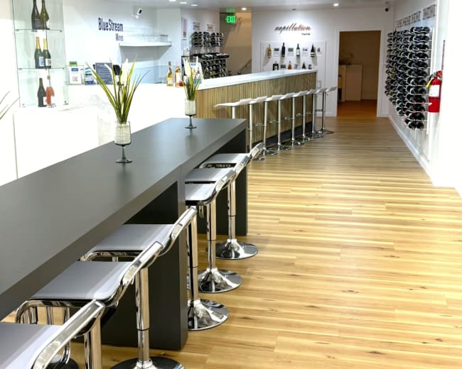 The image shows a modern tasting room with a long bar, high stools, and shelves displaying wine bottles along the walls.