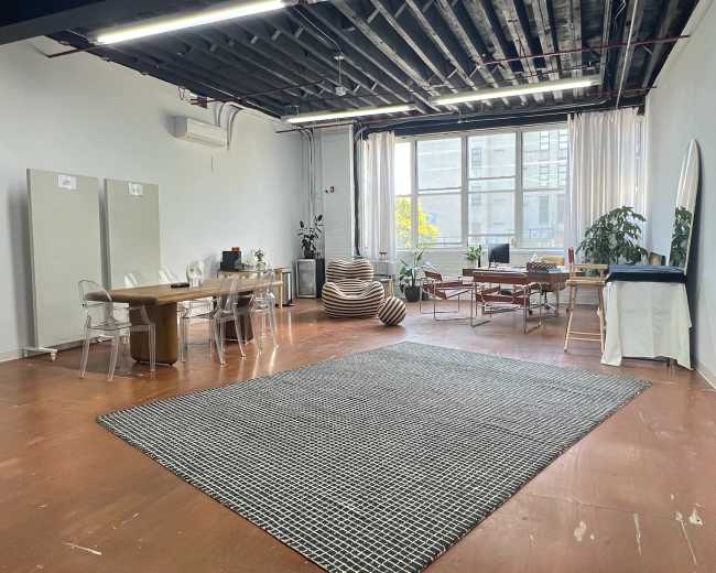 The image shows a spacious, well-lit studio with wooden floors, a large area rug, a dining table, and various seating arrangements.
