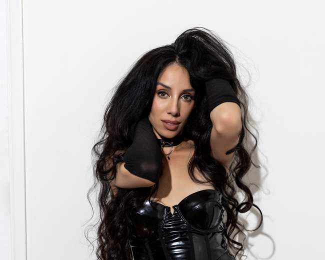 A woman with long, curly hair poses confidently in a black, shiny dress against a plain white wall.