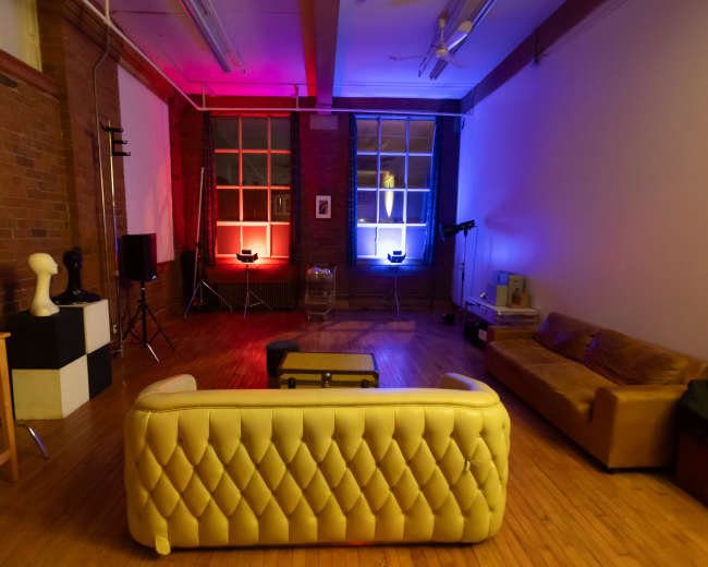 The image shows a spacious room with brick walls, featuring a yellow leather couch, a coffee table, and various lighting equipment arranged around two windows illuminated in red and blue.