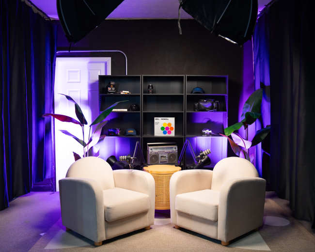 The image shows a cozy studio setup with two light-colored chairs facing each other in front of a shelf displaying various items, illuminated by purple lighting.