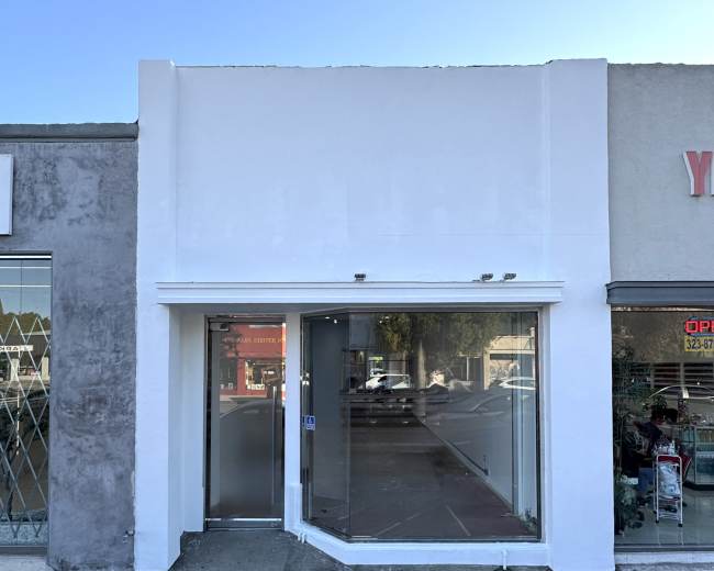 The image shows a vacant storefront with a large glass window and a white facade, situated between two neighboring businesses.