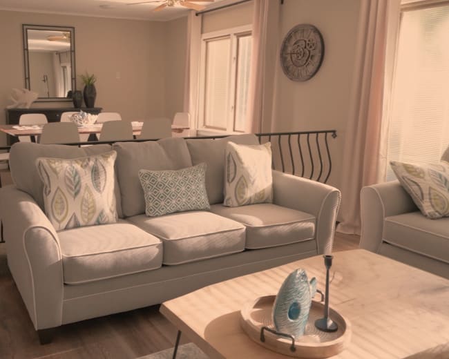 The image shows a well-lit living area featuring a light gray sofa with decorative pillows, a round coffee table, and a dining area in the background.