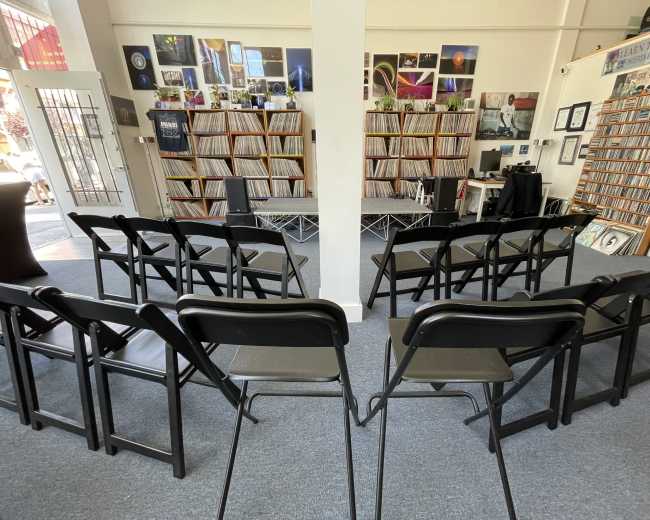 The image shows a room with rows of black chairs facing a small stage, surrounded by shelves filled with records and framed photographs.