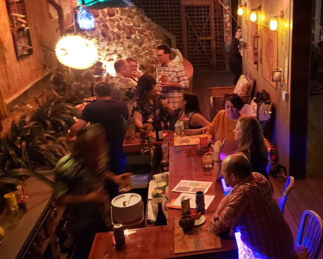 A lively bar scene with patrons socializing, some seated at the bar while others engage in conversation in a dimly lit outdoor area.