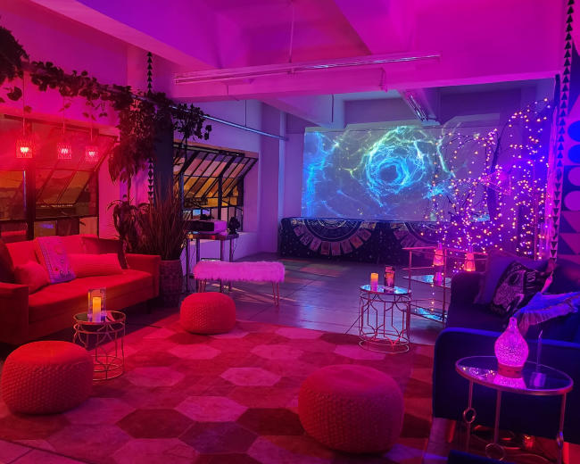 The image shows a vibrant lounge space with colorful lighting, a projector screen displaying swirling visuals, and various seating options including sofas and poufs.