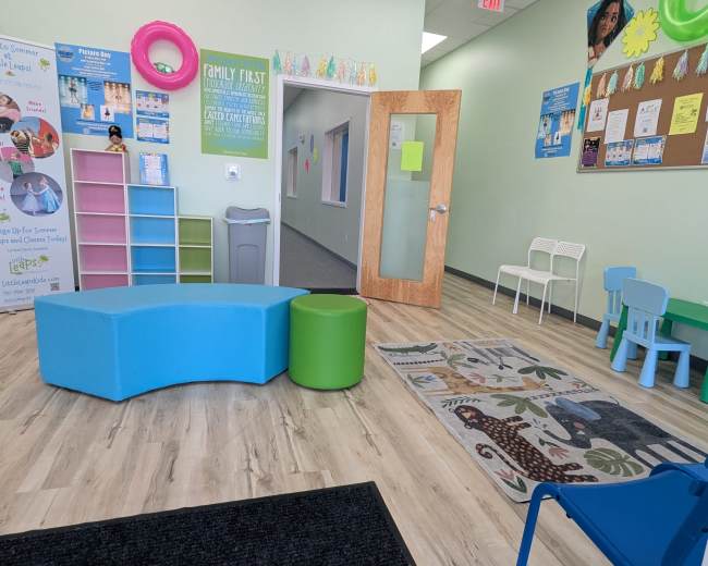 The image shows a bright waiting area with colorful furniture, including a blue curved couch, a green stool, and small tables and chairs designed for children.