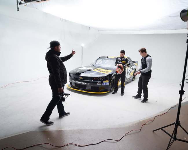 A car model is positioned in a studio while two individuals, one holding a helmet, interact with a photographer.