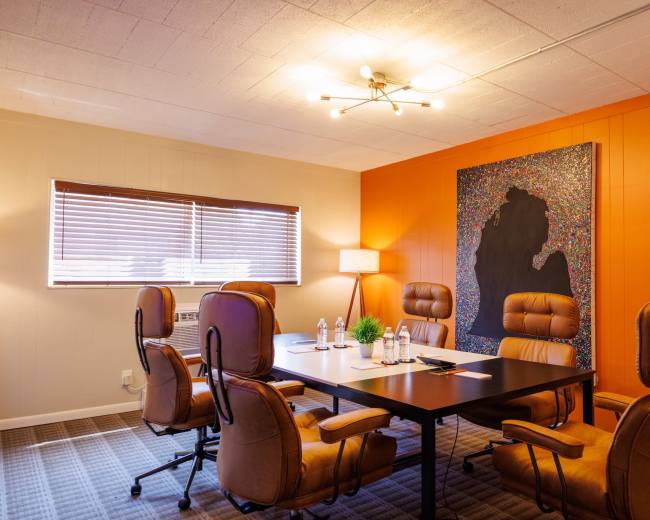 The image shows a modern conference room with a large table surrounded by four brown chairs, a lamp, a window with blinds, and an artistic wall piece featuring vibrant colors against an orange wall.