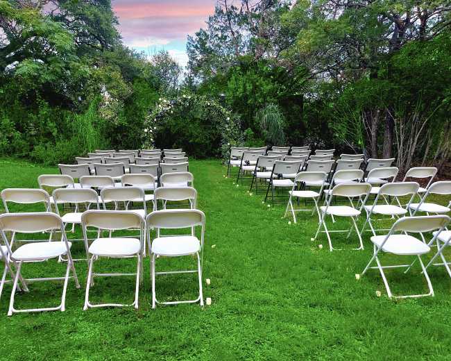 Rows of folding chairs are arranged on a grassy area surrounded by trees.