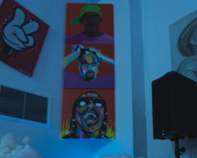 A candle sits on a coffee table in front of a sofa, with colorful artwork featuring faces on the wall behind it.