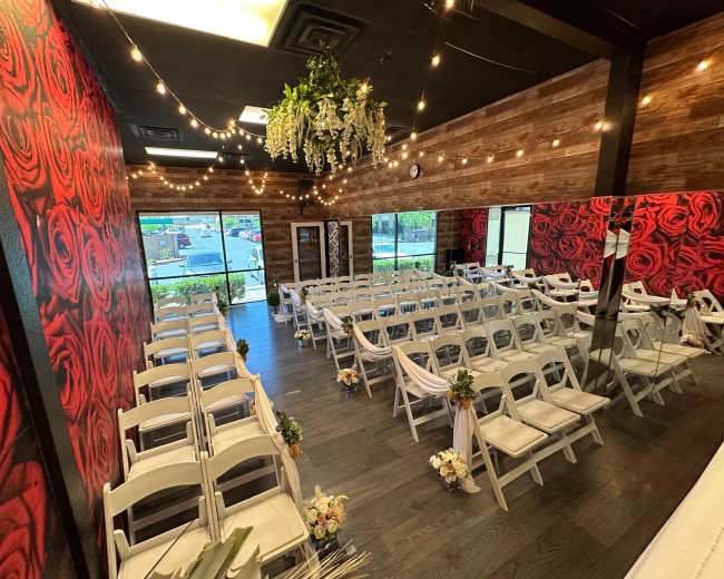 A wedding venue adorned with rose-covered walls, featuring rows of white chairs set up for a ceremony under string lights and decorative floral arrangements.