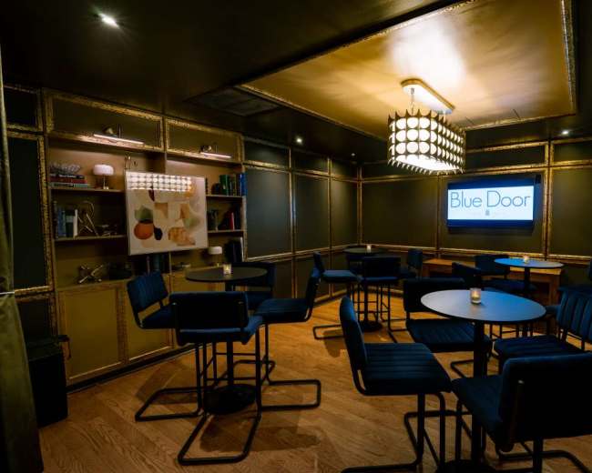 The image shows a dimly lit interior of a lounge or bar named "Blue Door," featuring several round tables and high-backed chairs against a backdrop of decorative shelves.