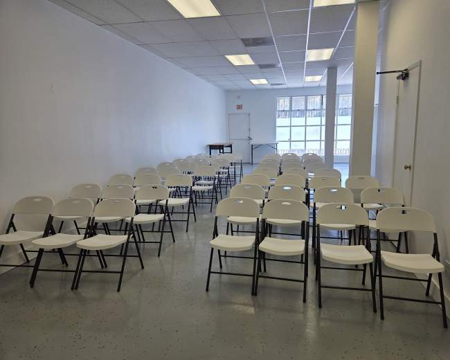 A row of white folding chairs is arranged in neat alignment within a sparsely furnished, well-lit room.