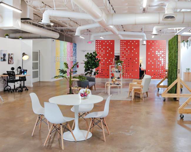 The image shows a modern open office space with a round table and chairs, colorful wall decor, plants, and various workstations.