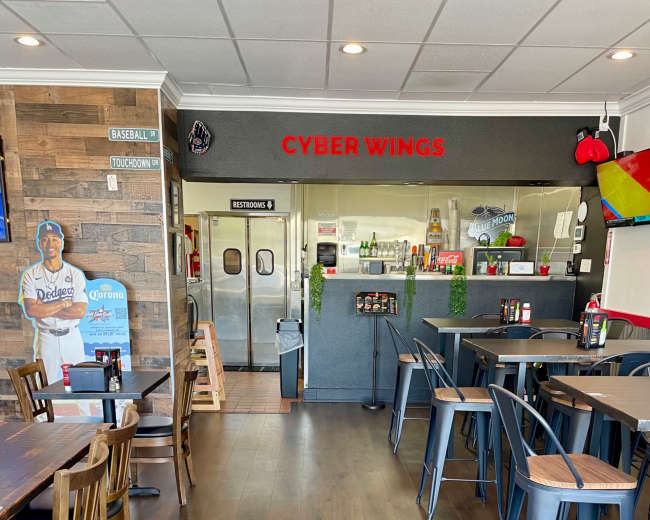 The image shows the interior of a restaurant named "Cyber Wings," featuring wooden tables, metal chairs, and a visible kitchen area in the background.