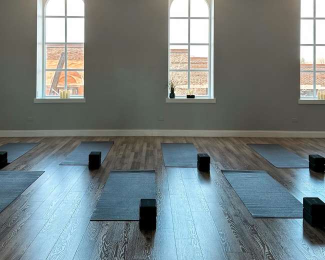 The image shows a spacious room with yoga mats and blocks arranged in parallel rows, illuminated by natural light from large windows.