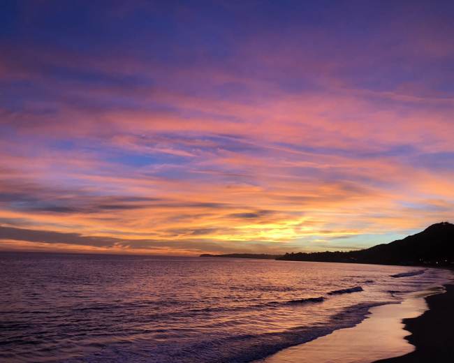 The image shows a beach at sunset, with vibrant colors in the sky reflecting on the water.