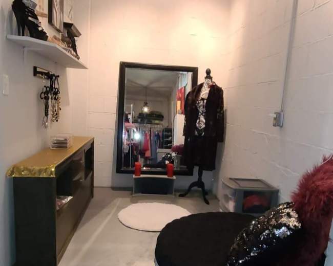 A narrow room featuring a large mirror, a black circular seating area, a wooden shelf with clothes, and several lights above.