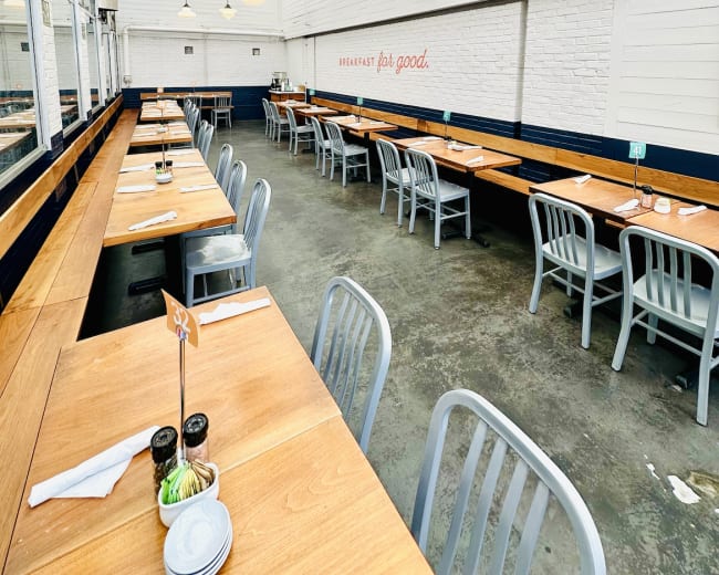 The image shows a spacious restaurant interior with long wooden tables and metal chairs arranged under a glass ceiling.