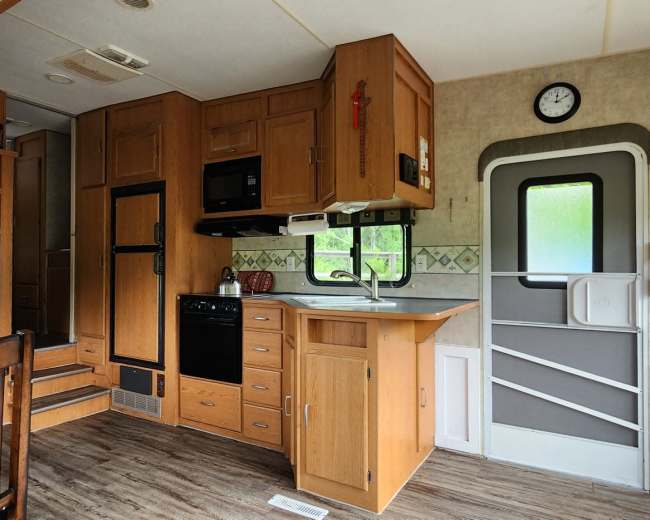 The image shows the interior of a camper with wooden cabinetry, a kitchen area, and a dining table.