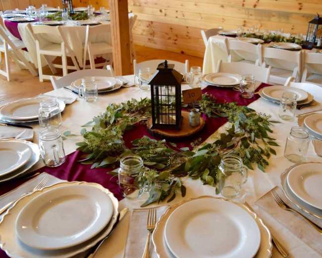 A round dining table is set with white plates and glass jars, surrounded by a greenery centerpiece and a lantern, against a backdrop of wooden walls and additional tables.