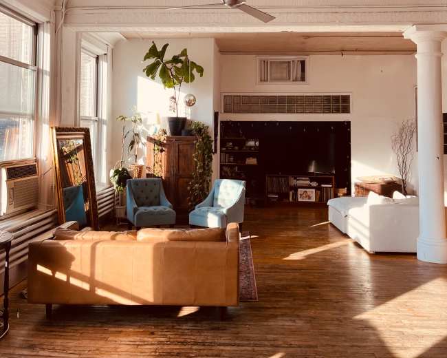 The image shows a well-lit, spacious living area featuring a mix of furniture, including a leather couch, blue chairs, and a white sofa, along with a large plant and decorative accents in a room with high ceilings and ornate details.