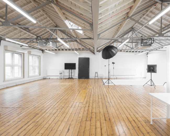 The image shows a spacious, well-lit studio with wooden floors, high ceilings, and various lighting equipment arranged throughout the room.