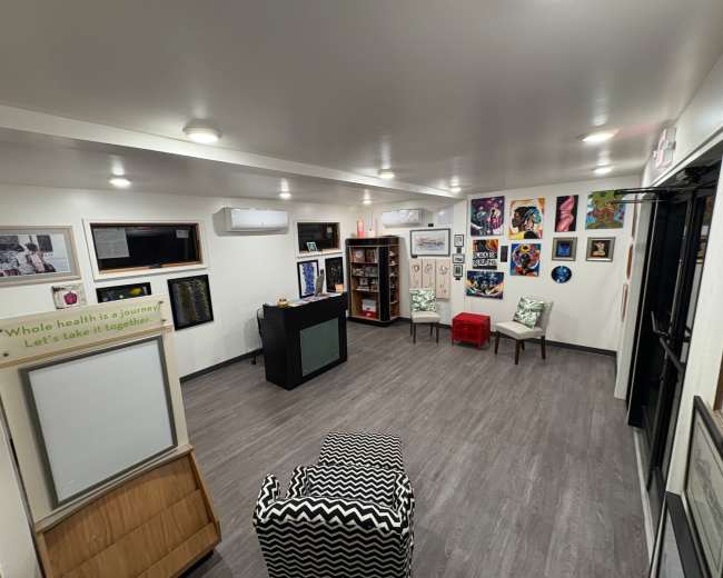 The image shows a brightly lit room featuring a variety of artworks on the walls, a seating area with patterned chairs, and a small display on one side.