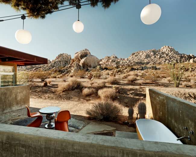 A modern outdoor space features a circular table with orange chairs, a freestanding bathtub, and boulders in the background under clear blue skies.