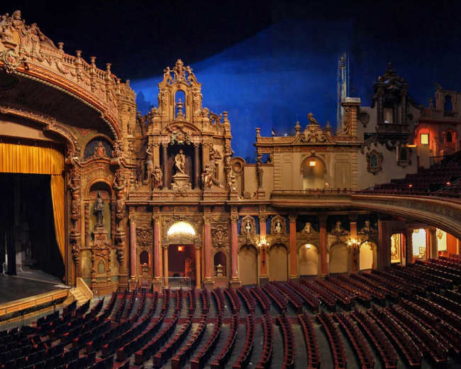 The image shows an ornate theater interior with a grand stage framed by intricate architectural details and a large, golden curtain.