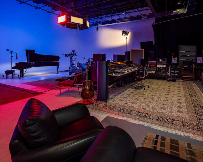 The image shows a music studio with a grand piano, recording equipment, and a comfortable seating area, all set against a backdrop of colored lighting.