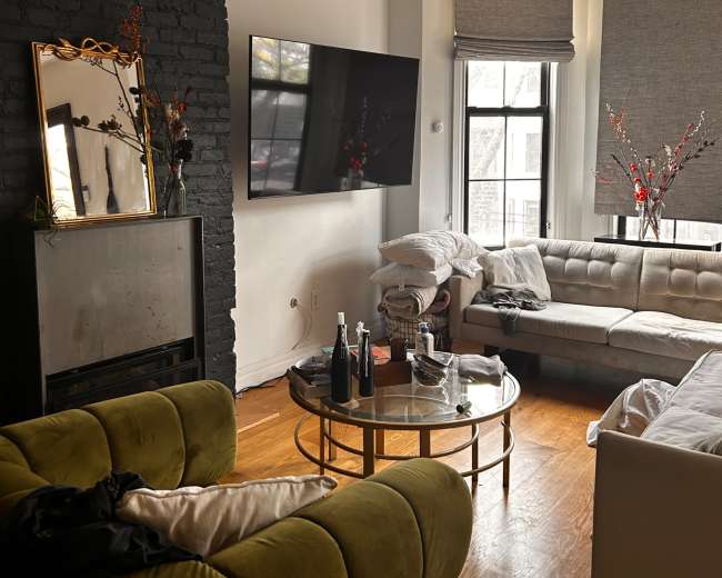 The image shows a modern living room featuring a green sofa, a round coffee table with drinks, a large wall-mounted TV, and large windows with shades.
