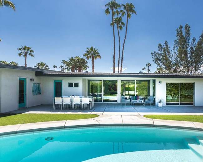 A modern backyard features a swimming pool with a seating area and dining set, surrounded by palm trees and a spacious patio.